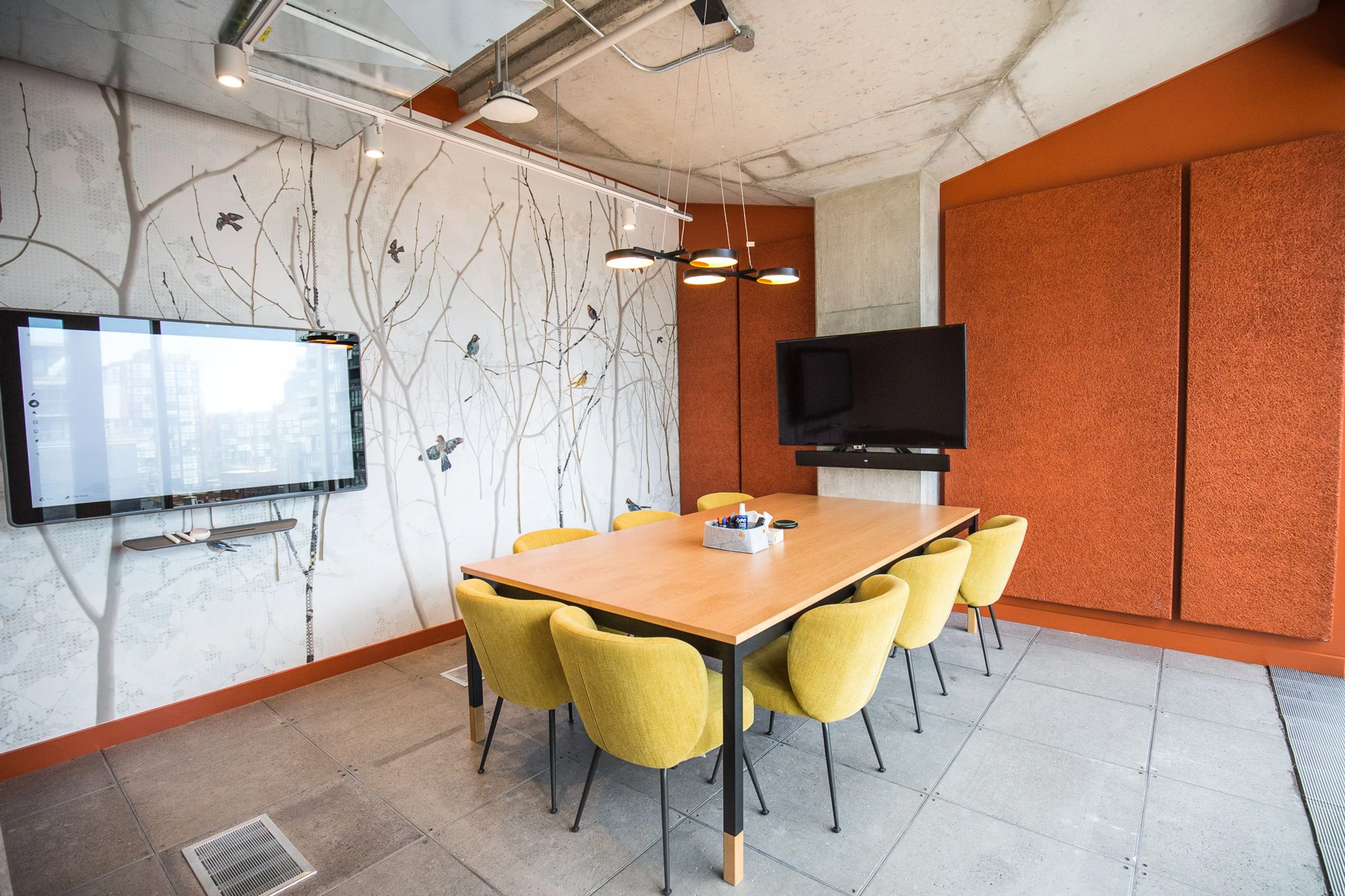 Meeting room with a bird-and-branch mural, exposed concrete, burnt-orange acoustic felt and a warm-oak table